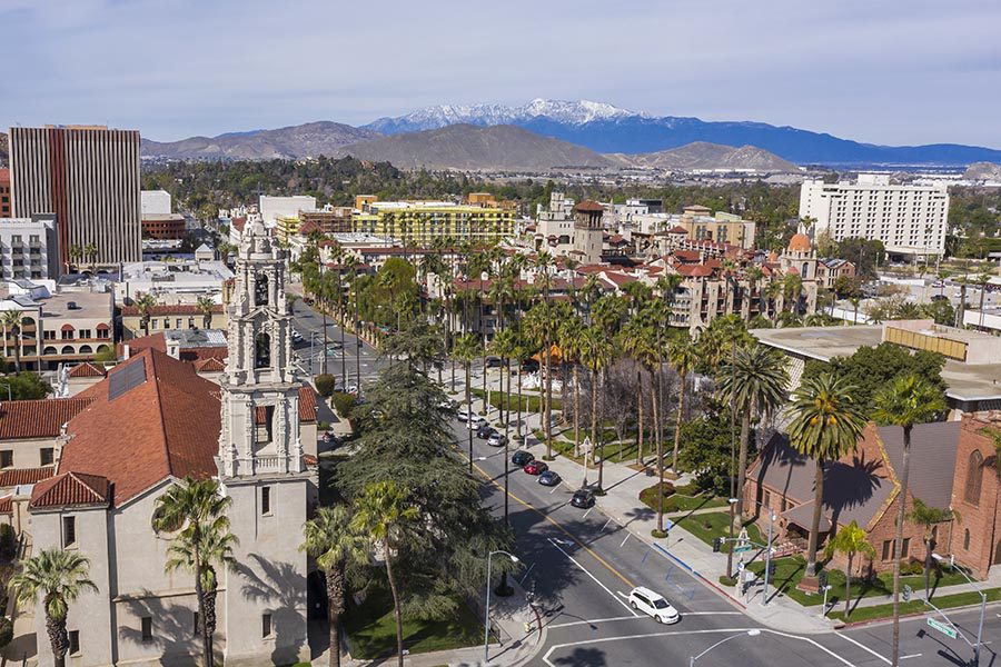 About Our Agency - Riverside, California Seen From Above, A Street Lined With Palm Trees, Large Buildings, and Snow-Capped Mountains in the Distance
