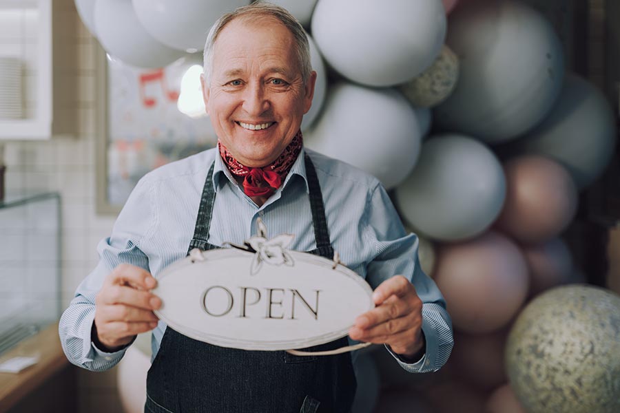 Business Insurance - Business Owner Wearing Overalls and a Red Bandana Holds an Open Sign, A Balloon Arch Behind Him