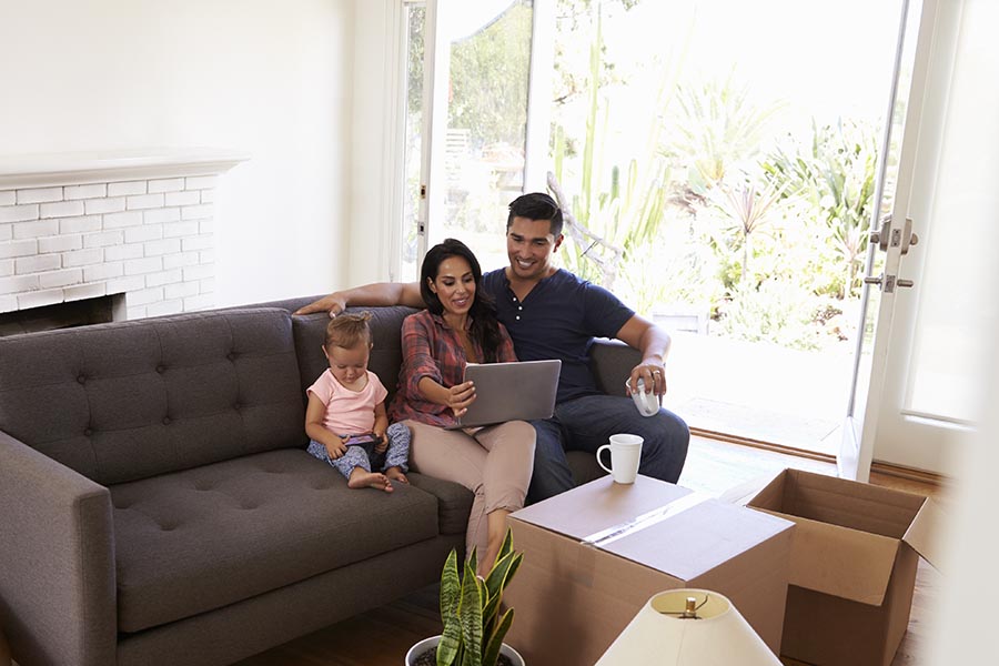 Client Center - A Couple and Their Young Daughter Use a Computer on a Gray Couch, Moving Boxes All Around Them
