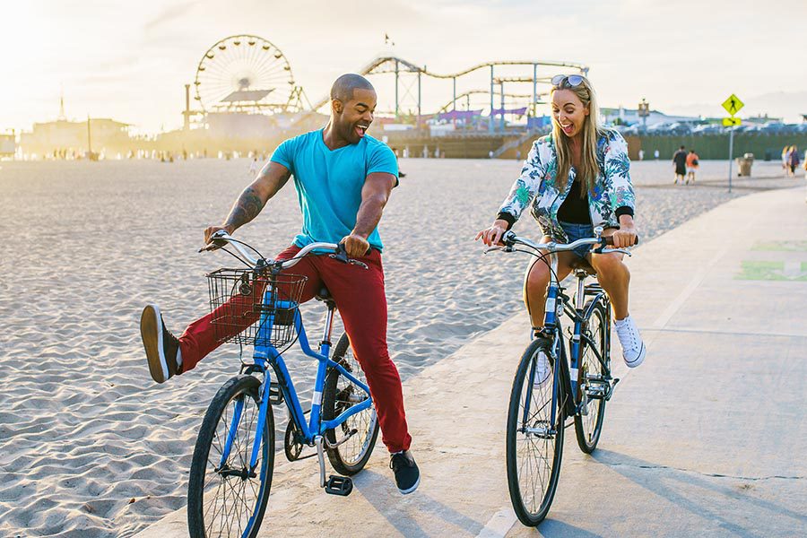 Personal Insurance - Happy Couple Laughing and Riding Bikes on a Beach Path, An Amusement Park Behind Them